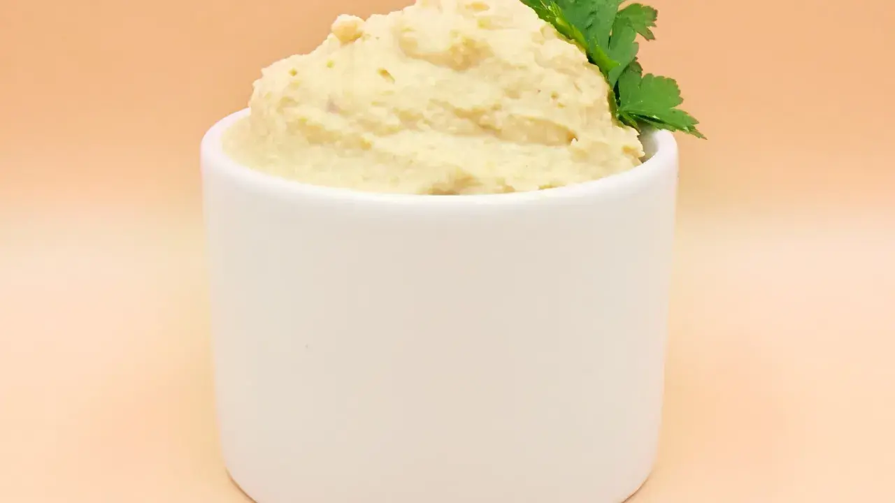 Kremowy hummus w białej miseczce, ozdobiony gałązką pietruszki.