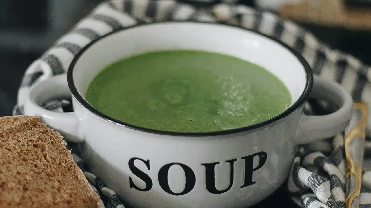 Zielony kremowy zupa w białej misce z napisem "SOUP", obok kromka chleba. Idealne rozwiązanie, jak mrozić zupy na później.