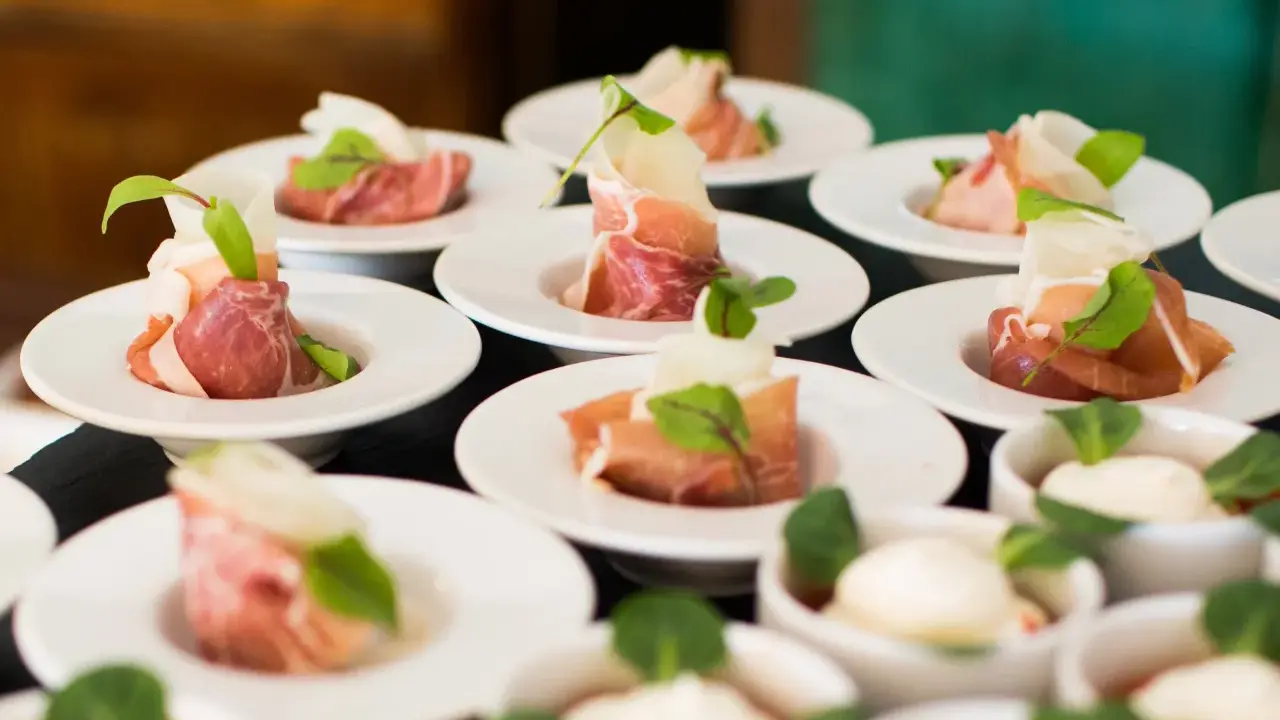Eleganckie przekąski na białych talerzykach, zawinięte w szynkę parmeńską z listkami ziół. To właśnie catering, który zachwyca smakiem i wyglądem.