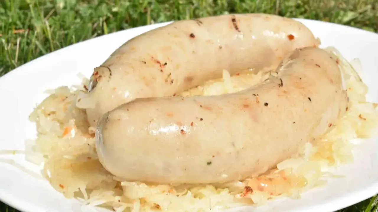Dwie białe kiełbaski, idealnie przyrumienione na grillu, leżą na talerzu z kiszoną kapustą. Pyszna kiszka ziemniaczana z grilla w letnim otoczeniu.