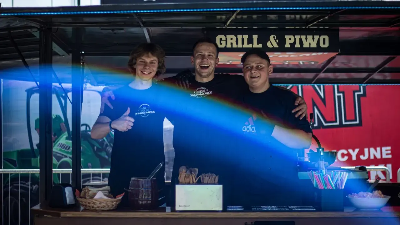 Trzech uśmiechniętych mężczyzn za ladą cateringową z napisem "Grill & Piwo".