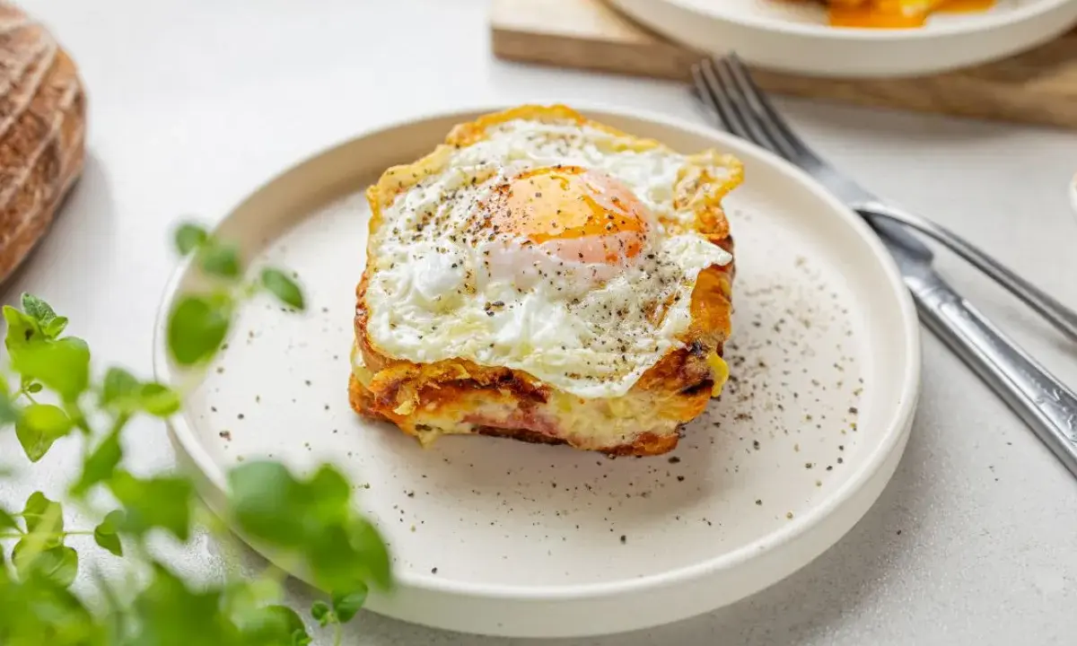 Pyszny croque madame na talerzu, z jajkiem sadzonym na wierzchu i pieprzem. Obok widoczny widelec i nóż.