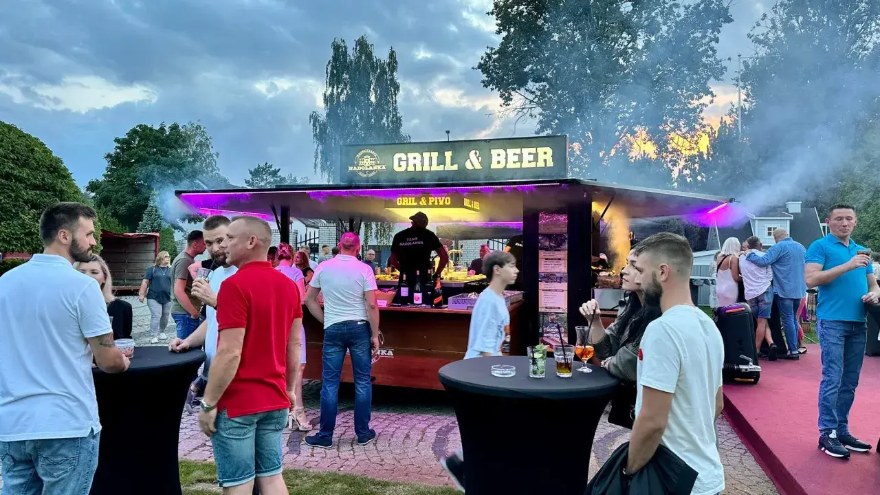 Ludzie przy stoisku z grillem i piwem, z dymem unoszącym się w powietrzu.