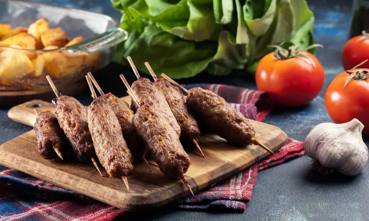 Aksamitne cevapi na drewnianej desce, obok pomidory, czosnek i pieczone ziemniaki.