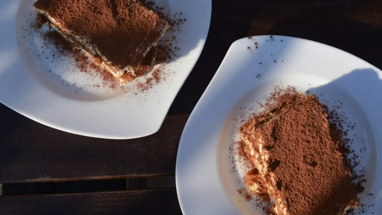 Dwa kawałki tiramisu posypane kakao na białych talerzach. Idealne do mocnej kawy, która podkreśli smak.