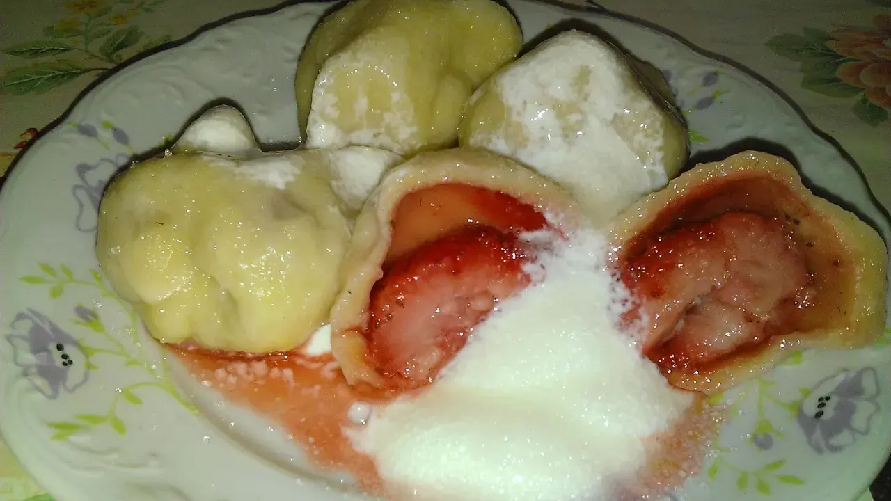 Jak gotować pierogi mrożone z truskawkami? Na talerzu pyszne, gorące pierogi z owocami, polane śmietaną i cukrem.
