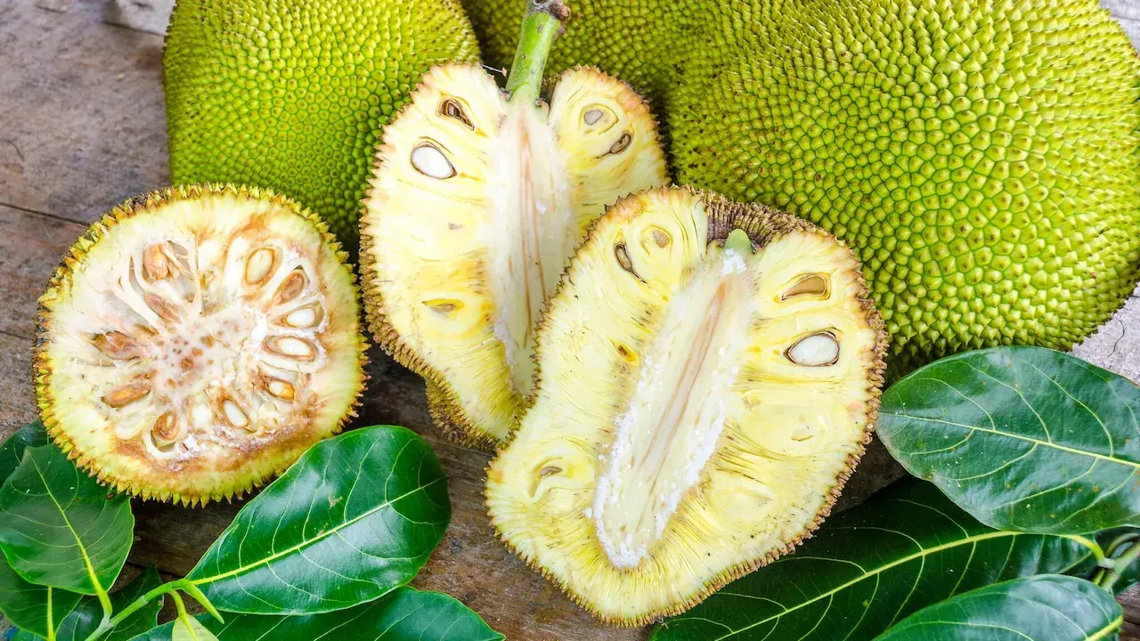 Cały i przekrojony chlebowiec (jackfruit) z widocznymi nasionami i liśćmi na drewnianym tle.