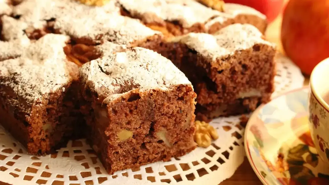 Kawałki aromatycznego ciasta cynamonowego z orzechami, posypane cukrem pudrem, obok jabłek i filiżanki herbaty.