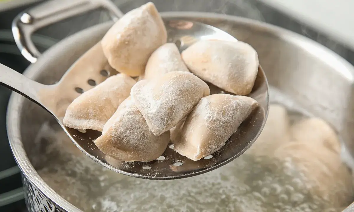 Mrożone pierogi na łyżce cedzakowej nad gotującą się wodą.
