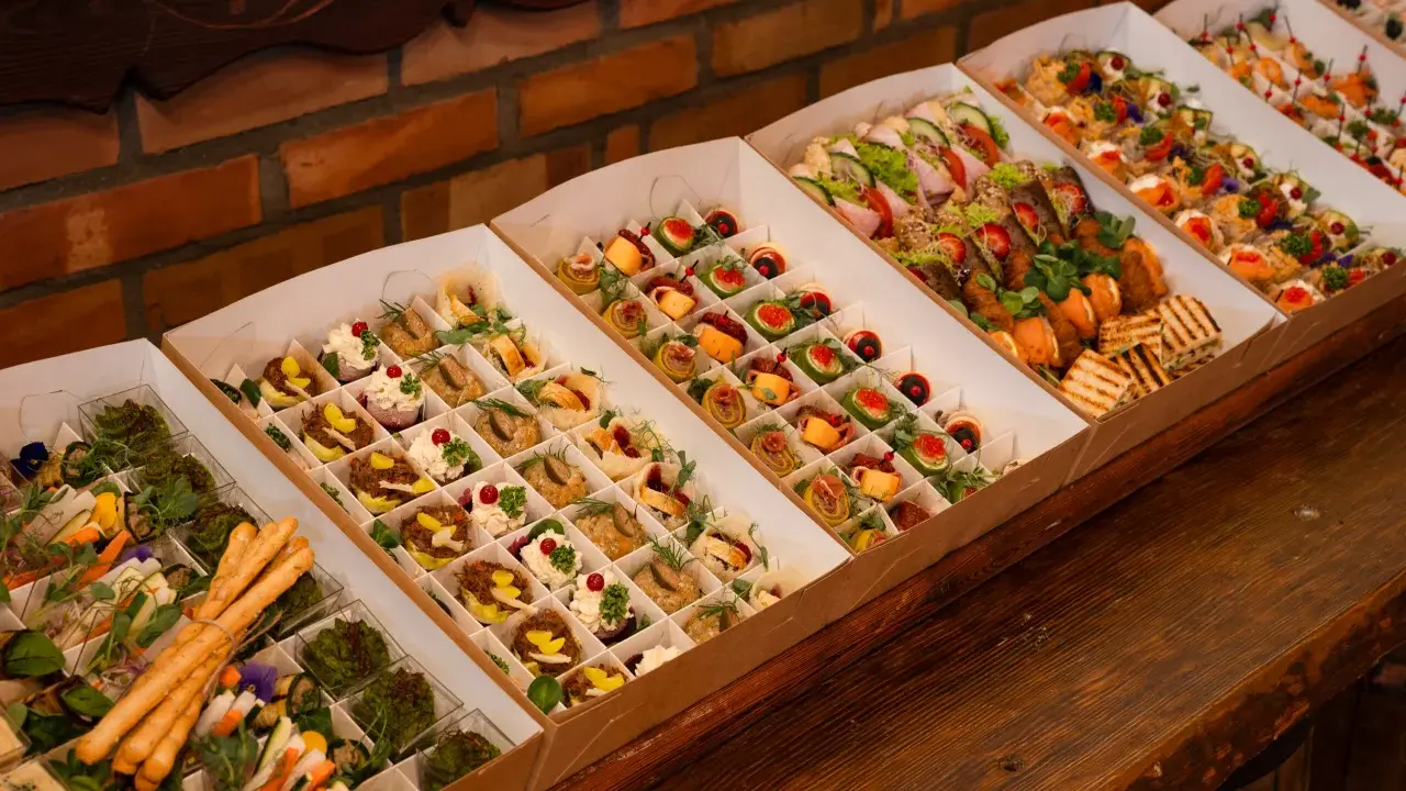 Elegancki catering do domu: pięknie ułożone przekąski w kartonowych pudełkach, od mini kanapek po rolki z łososiem i warzywami.