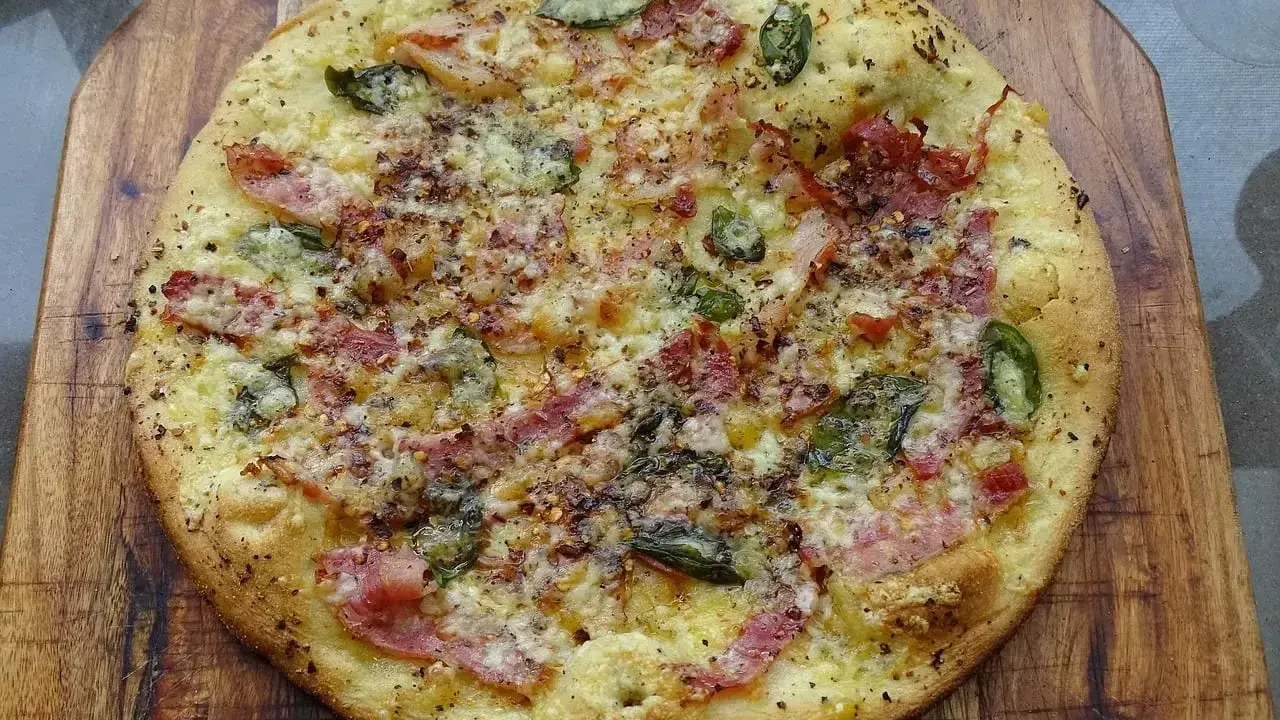Pyszna pizza bianca z szynką, serem i zielonymi dodatkami, posypana przyprawami, na drewnianej desce.