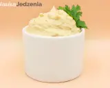 Kremowy hummus w białej miseczce, ozdobiony gałązką pietruszki.