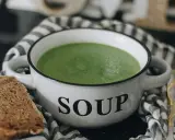 Zielony kremowy zupa w białej misce z napisem "SOUP", obok kromka chleba. Idealne rozwiązanie, jak mrozić zupy na później.