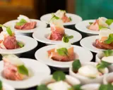 Eleganckie przekąski na białych talerzykach, zawinięte w szynkę parmeńską z listkami ziół. To właśnie catering, który zachwyca smakiem i wyglądem.