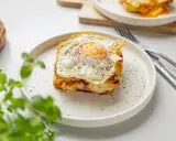 Pyszny croque madame na talerzu, z jajkiem sadzonym na wierzchu i pieprzem. Obok widoczny widelec i nóż.