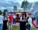 Ludzie przy stoisku z grillem i piwem, z dymem unoszącym się w powietrzu.