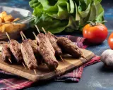 Aksamitne cevapi na drewnianej desce, obok pomidory, czosnek i pieczone ziemniaki.