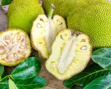 Cały i przekrojony chlebowiec (jackfruit) z widocznymi nasionami i liśćmi na drewnianym tle.