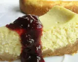 Kawałek kremowego sernika z polewą z ciemnych owoców na białym talerzu.