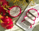 Dwie całe i jedna przekrojona pitahaya czerwona, z której wyjęto kilka kawałków. Owoce leżą na drewnianym blacie, obok żółte kwiaty.