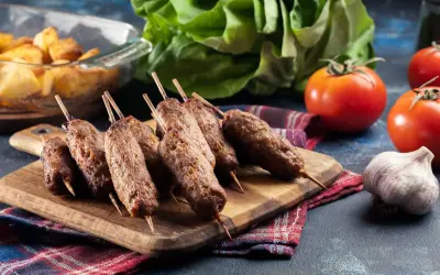 Aksamitne cevapi na drewnianej desce, obok pomidory, czosnek i pieczone ziemniaki.