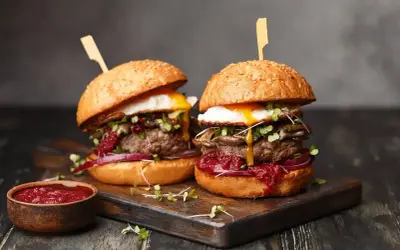 Dwa soczyste burgery z grilla, z jajkiem sadzonym i dodatkami, prezentują się apetycznie na desce.