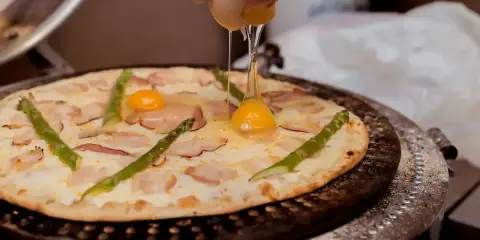 Pizza z jajkiem i szparagami, na którą wylewane jest surowe jajko.