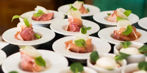 Eleganckie przekąski na białych talerzykach, zawinięte w szynkę parmeńską z listkami ziół. To właśnie catering, który zachwyca smakiem i wyglądem.