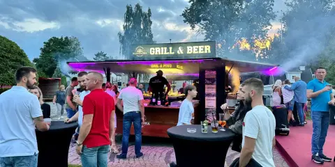 Ludzie przy stoisku z grillem i piwem, z dymem unoszącym się w powietrzu.