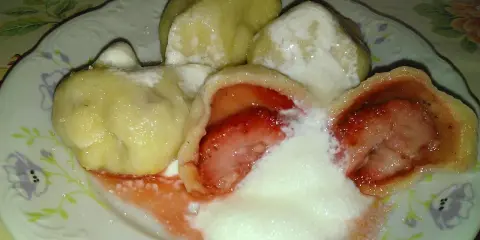 Jak gotować pierogi mrożone z truskawkami? Na talerzu pyszne, gorące pierogi z owocami, polane śmietaną i cukrem.