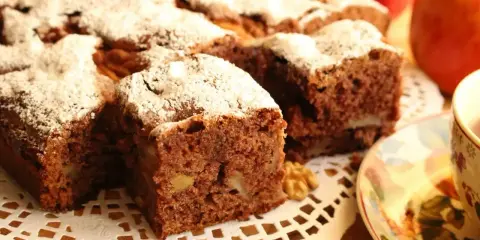 Kawałki aromatycznego ciasta cynamonowego z orzechami, posypane cukrem pudrem, obok jabłek i filiżanki herbaty.