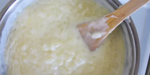 Kremowa zupa ryżanka gotuje się w garnku, drewniana łyżka miesza gęsty, biały płyn z widocznymi ziarenkami ryżu.
