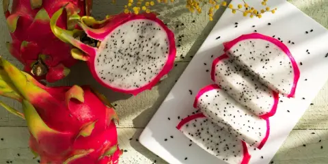 Dwie całe i jedna przekrojona pitahaya czerwona, z której wyjęto kilka kawałków. Owoce leżą na drewnianym blacie, obok żółte kwiaty.