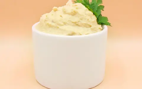 Kremowy hummus w białej miseczce, ozdobiony gałązką pietruszki.