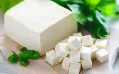 Kostka tofu i pokrojone w kostkę tofu na desce, ozdobione zielonymi liśćmi.