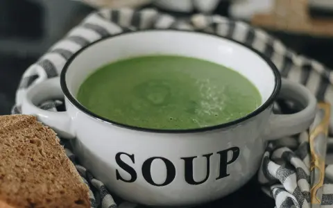 Zielony kremowy zupa w białej misce z napisem "SOUP", obok kromka chleba. Idealne rozwiązanie, jak mrozić zupy na później.