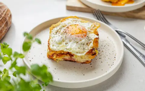Pyszny croque madame na talerzu, z jajkiem sadzonym na wierzchu i pieprzem. Obok widoczny widelec i nóż.