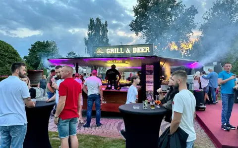 Ludzie przy stoisku z grillem i piwem, z dymem unoszącym się w powietrzu.