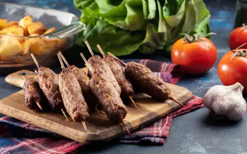 Aksamitne cevapi na drewnianej desce, obok pomidory, czosnek i pieczone ziemniaki.