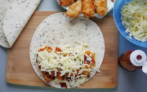 Tortilla z kurczakiem, serem i sosem na desce. Obok miska z serem i butelka ketchupu.