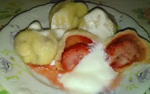 Jak gotować pierogi mrożone z truskawkami? Na talerzu pyszne, gorące pierogi z owocami, polane śmietaną i cukrem.