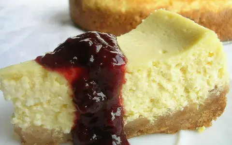 Kawałek kremowego sernika z polewą z ciemnych owoców na białym talerzu.