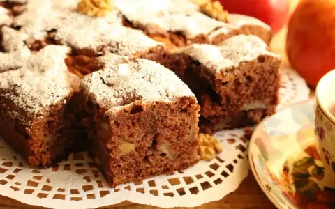 Kawałki aromatycznego ciasta cynamonowego z orzechami, posypane cukrem pudrem, obok jabłek i filiżanki herbaty.