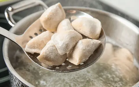 Mrożone pierogi na łyżce cedzakowej nad gotującą się wodą.