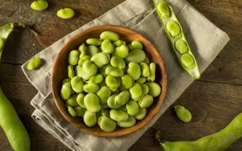 Miseczka pełna świeżych, zielonych fasolek fava, obok łuszczyn i pojedynczych ziaren na drewnianym blacie.