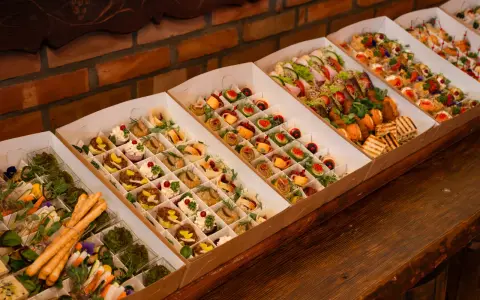 Elegancki catering do domu: pięknie ułożone przekąski w kartonowych pudełkach, od mini kanapek po rolki z łososiem i warzywami.