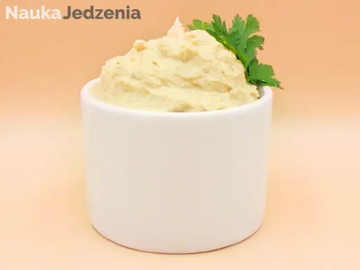 Kremowy hummus w białej miseczce, ozdobiony gałązką pietruszki.
