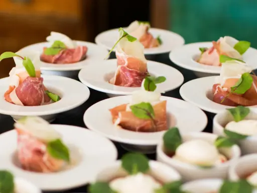 Eleganckie przekąski na białych talerzykach, zawinięte w szynkę parmeńską z listkami ziół. To właśnie catering, który zachwyca smakiem i wyglądem.