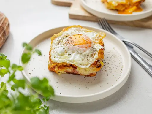 Pyszny croque madame na talerzu, z jajkiem sadzonym na wierzchu i pieprzem. Obok widoczny widelec i nóż.