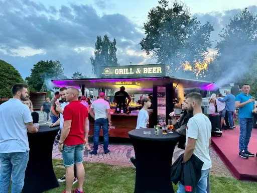 Ludzie przy stoisku z grillem i piwem, z dymem unoszącym się w powietrzu.